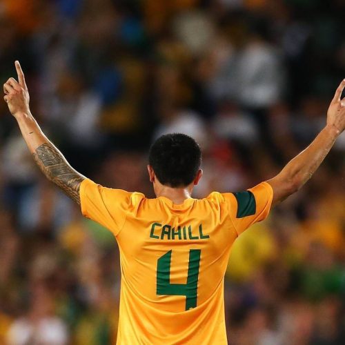 Jogador da seleção australiana de costas usando camisa amarela com detalhes verdes durante partida de futebol.
