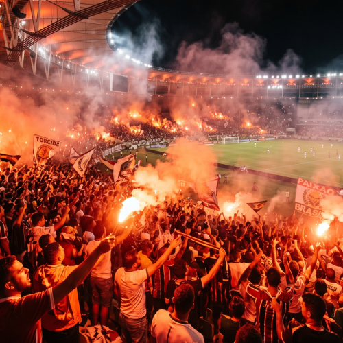 Fotografia realista e ampla de uma arquibancada de estádio de futebol lotada e iluminada por sinalizadores de fumaça vermelha e laranja, com torcedores vibrando intensamente. crie essa imagem