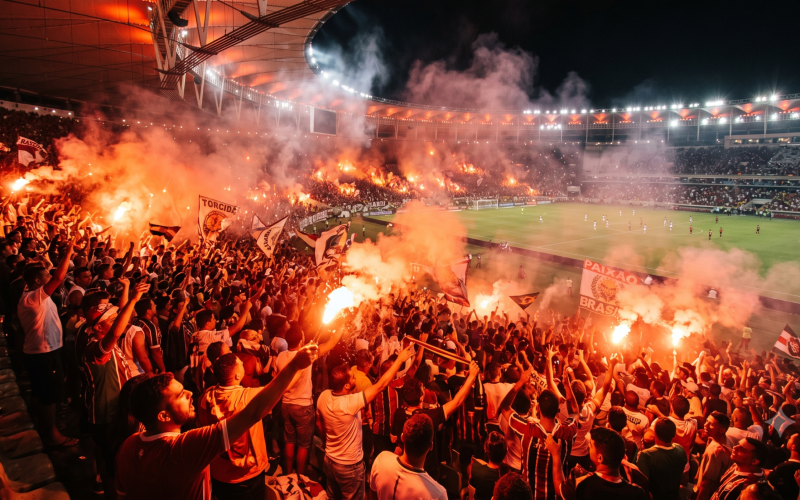 Fotografia realista e ampla de uma arquibancada de estádio de futebol lotada e iluminada por sinalizadores de fumaça vermelha e laranja, com torcedores vibrando intensamente. crie essa imagem