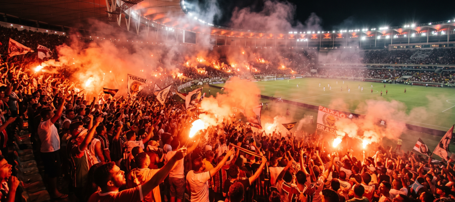 Fotografia realista e ampla de uma arquibancada de estádio de futebol lotada e iluminada por sinalizadores de fumaça vermelha e laranja, com torcedores vibrando intensamente. crie essa imagem